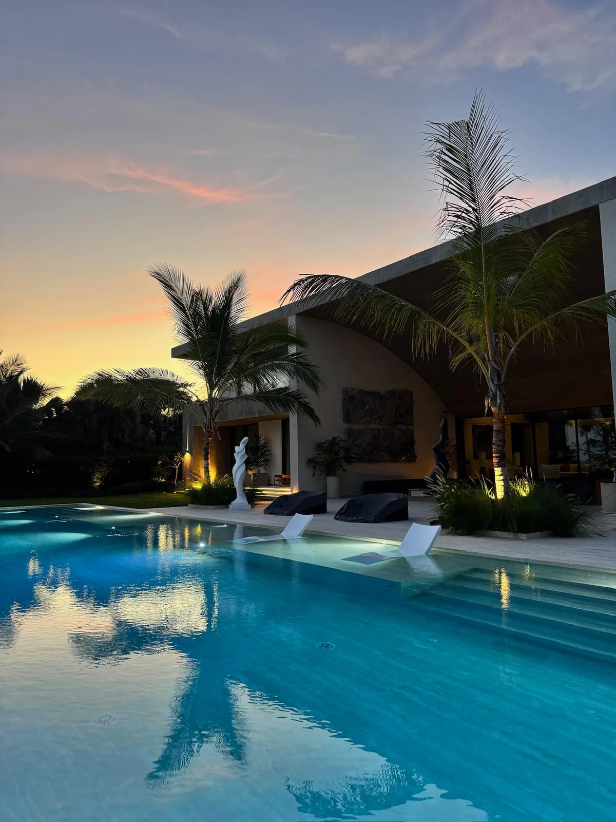 Arrecife estate at sunset, with an illuminated pool, uplit palms and a white sculpture on the terrace